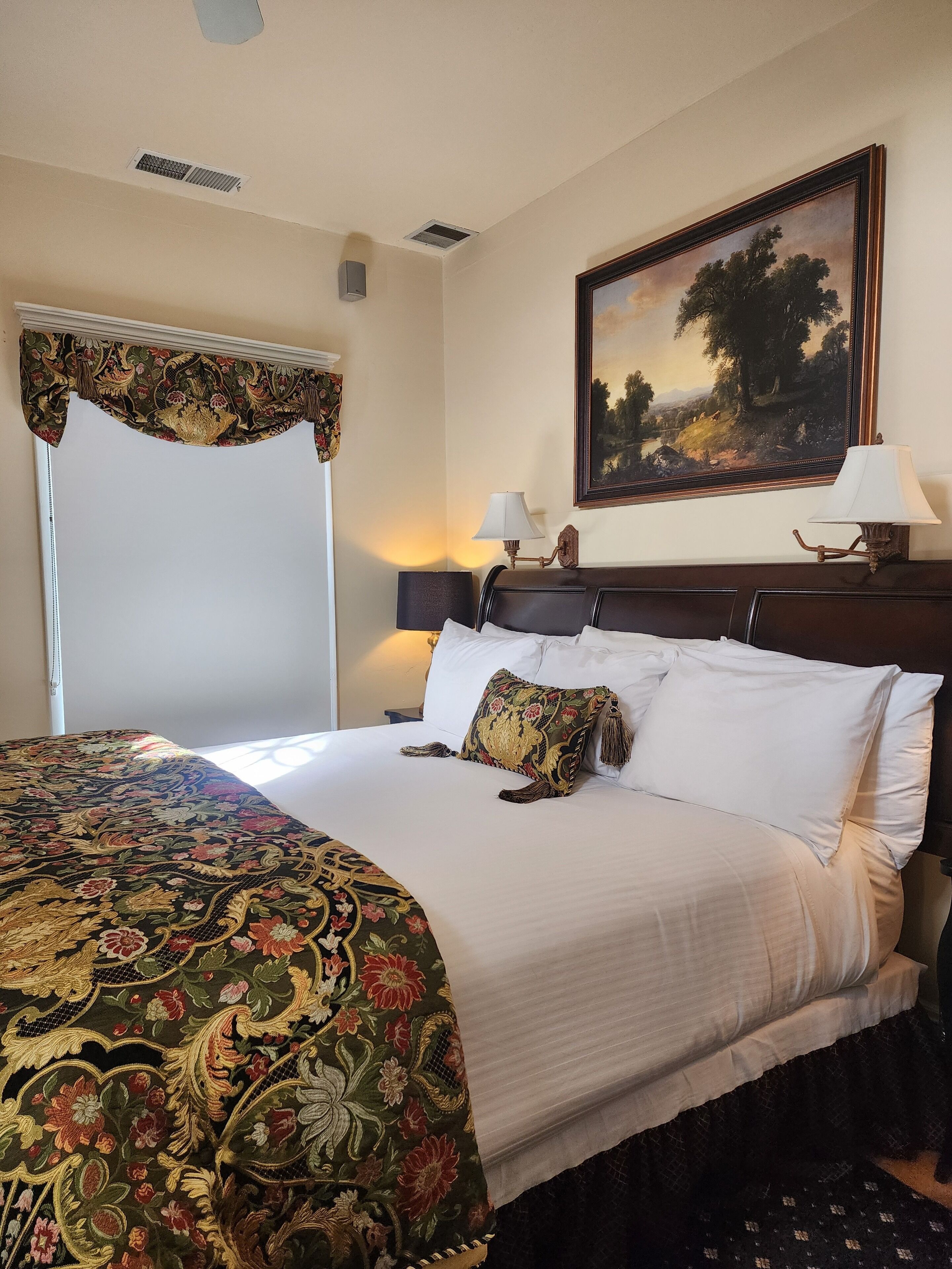 A neatly made bed with a floral bedspread, two bedside lamps, and a framed landscape painting on the wall.