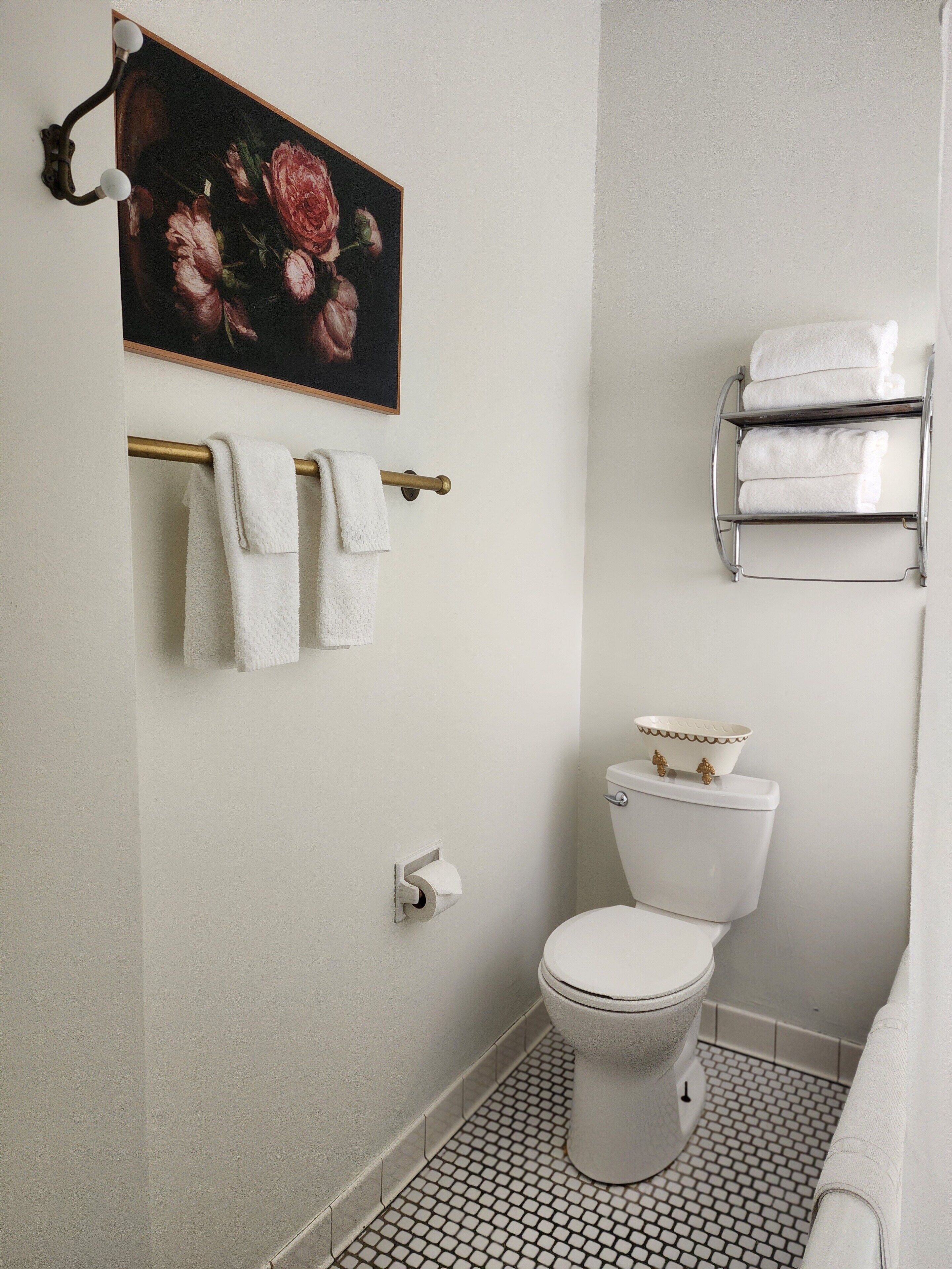 A bathroom with a toilet, a towel rack with towels, a painting above the towel rack, and a small shelf with a bowl.