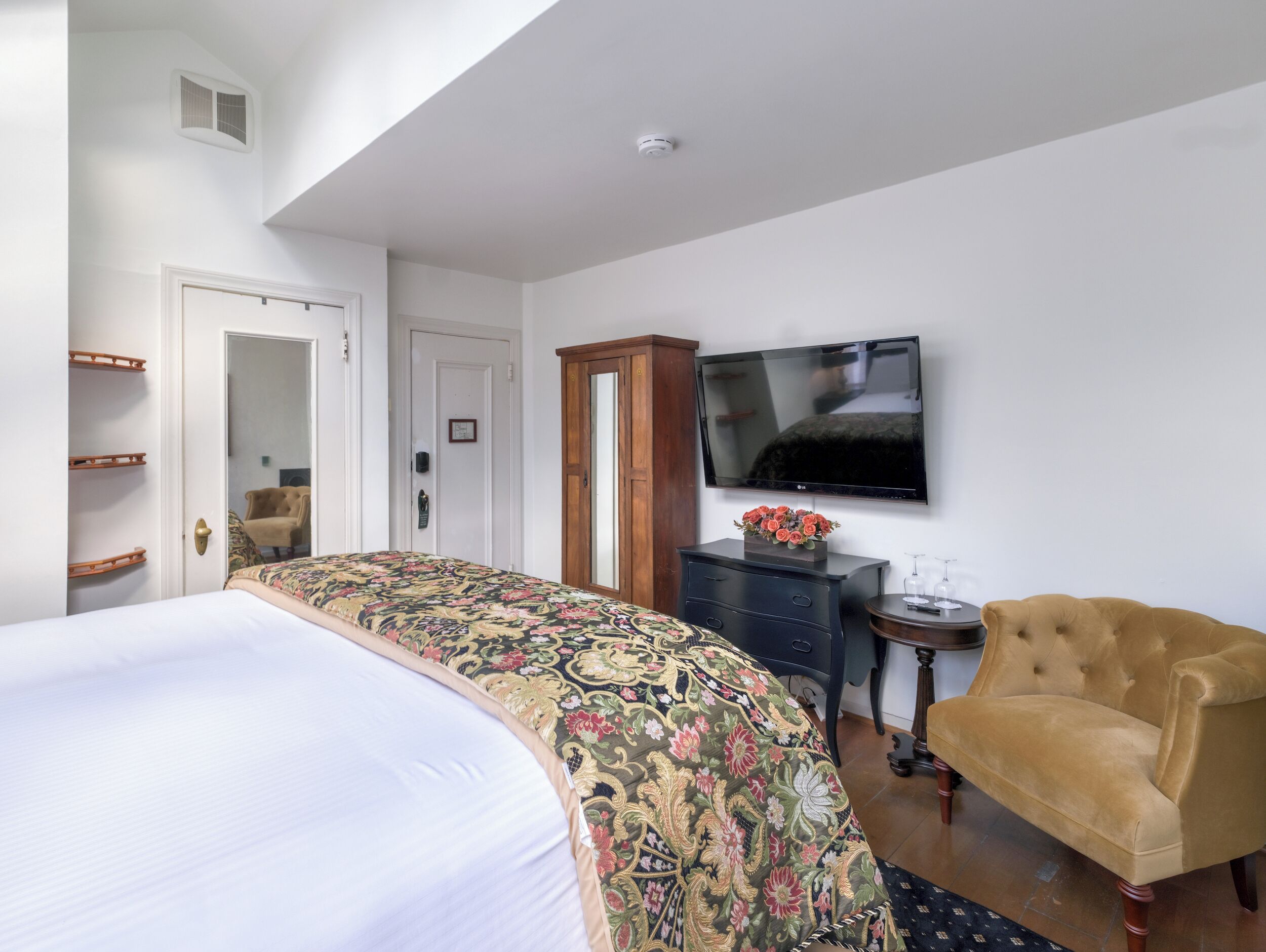 A hotel room with a large bed, a television mounted on the wall, a wooden wardrobe, a small table with a vase of flowers, and a plush armchair.