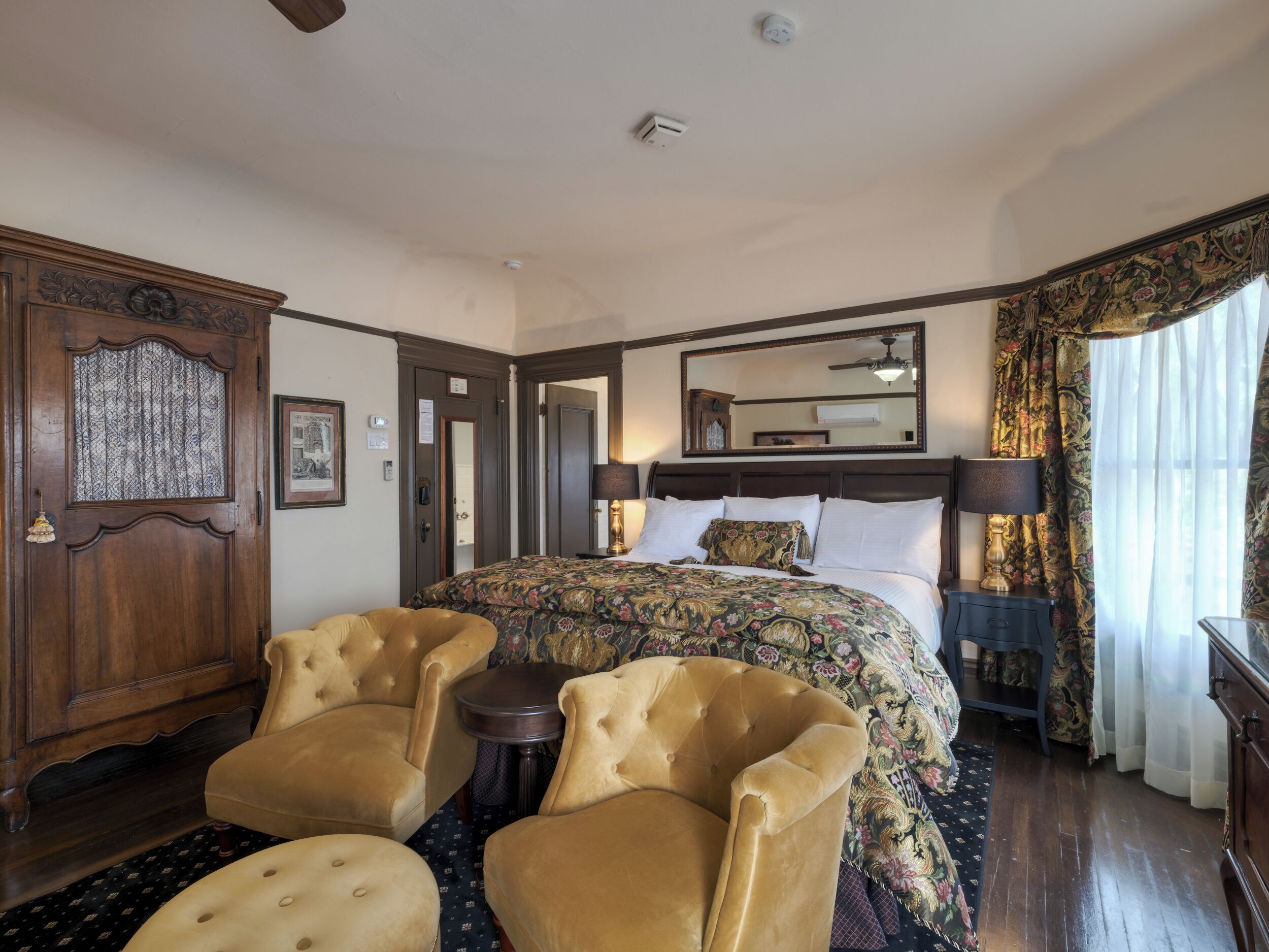 Une chambre d’hôtel avec un grand lit, deux fauteuils, une petite table, un miroir et un ventilateur de plafond.