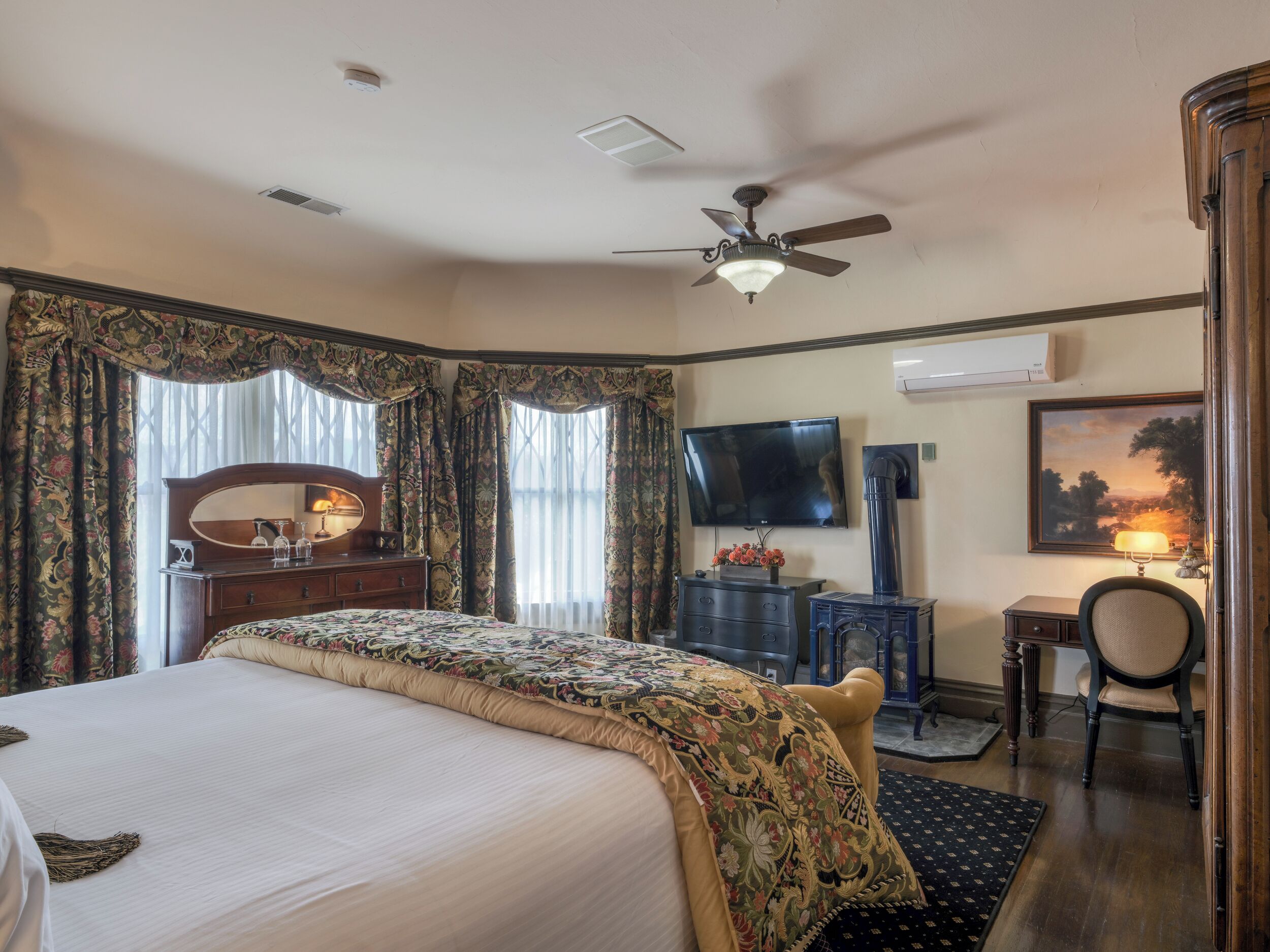 A hotel room with a large bed, a desk with a chair, a television, and a fireplace.