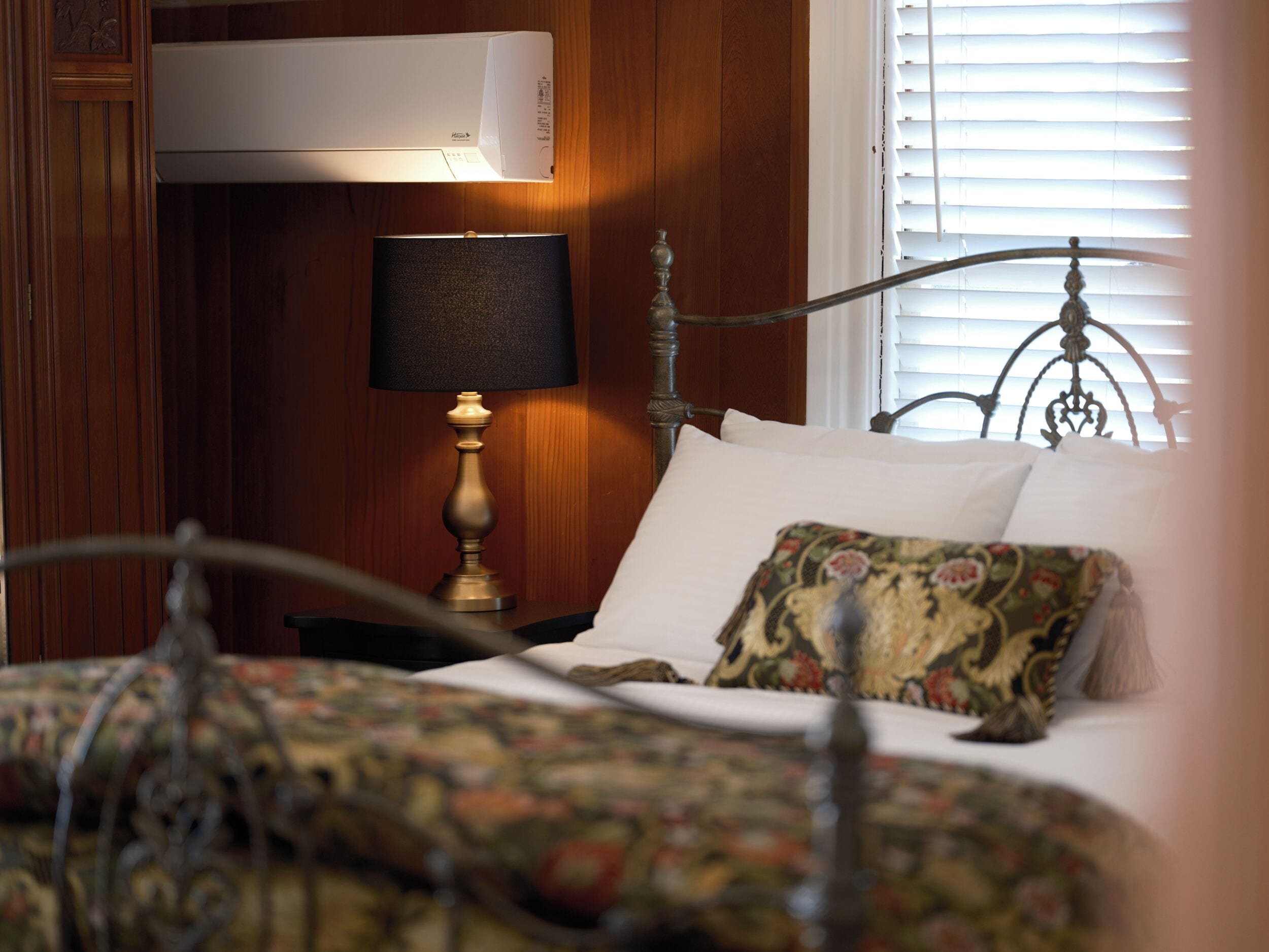A bedroom with a bed, a nightstand, a lamp, a window with blinds, and an air conditioning unit.