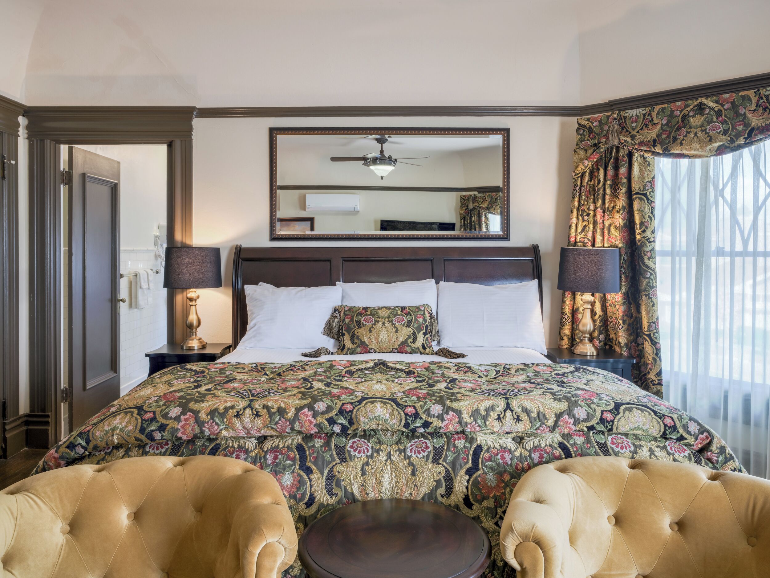 A hotel room with a large bed, two armchairs, a round coffee table, a ceiling fan, and a large mirror above the bed.