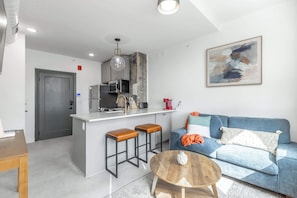 A modern kitchen with a central island, a dining area with stools, a sofa with cushions, and a coffee table.