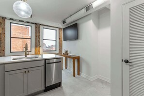 A modern kitchen with a stainless steel dishwasher, a wooden table with kitchenware, and a large window offering a city view.