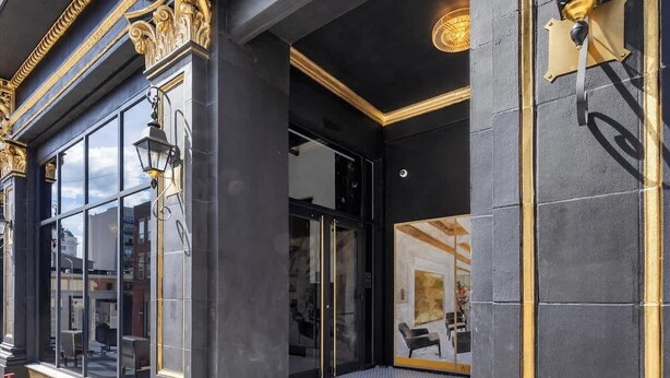 Une entrée d’immeuble avec une façade noire, des ornements décoratifs dorés et une porte en verre donnant sur l’intérieur.