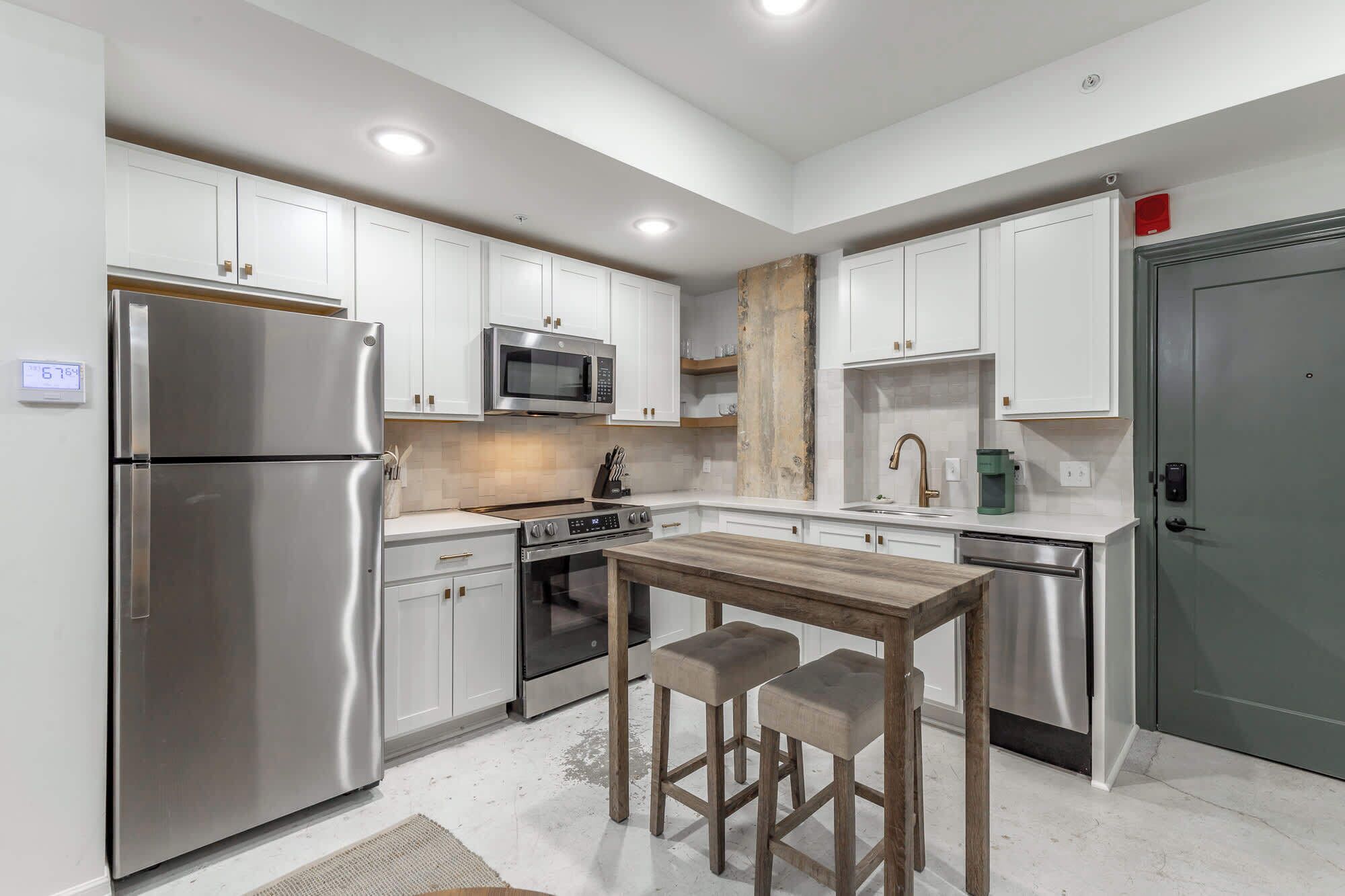 A modern kitchen with white cabinets, stainless steel appliances, a wooden table with stools, and a grey door.