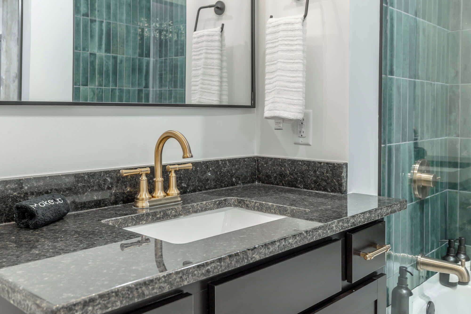 A bathroom with a granite countertop, a gold faucet, a white sink, and a glass shower enclosure.