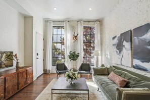 Ein Wohnzimmer mit einer Couch, Sesseln, einem Couchtisch und einem großen Fenster mit Vorhängen.