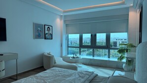 Un dormitorio moderno con una cama grande, un sofá, una mesita y un televisor. Hay dos cuadros enmarcados en la pared y una ventana con persianas.