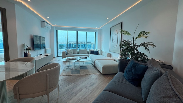 Una sala de estar moderna con sofá, mesa de centro y sillas. Se aprecia un amplio ventanal con vista a la ciudad.