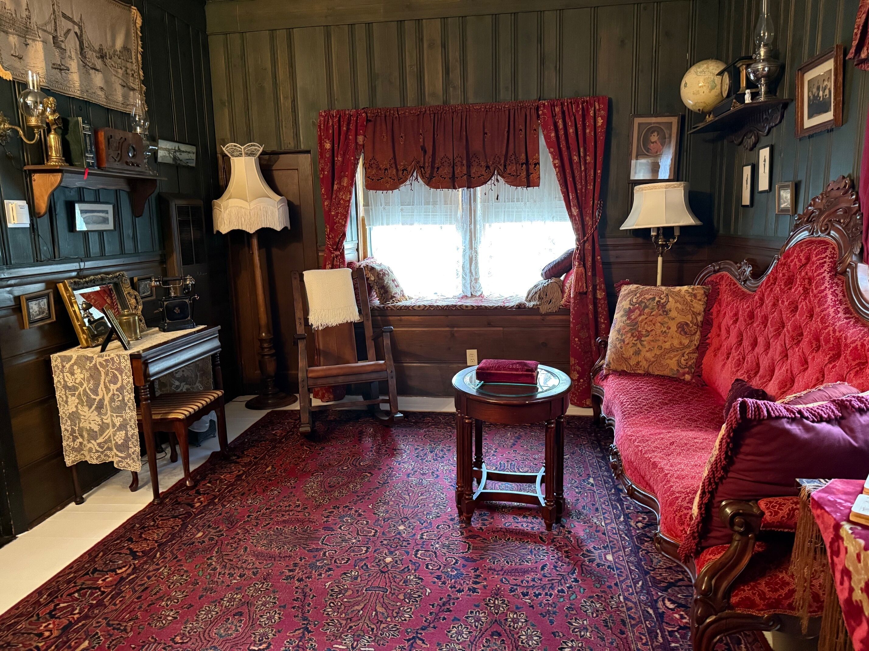 A traditional room with a red patterned carpet, a wooden sofa, a wooden chair, a small table, and a window with red curtains.