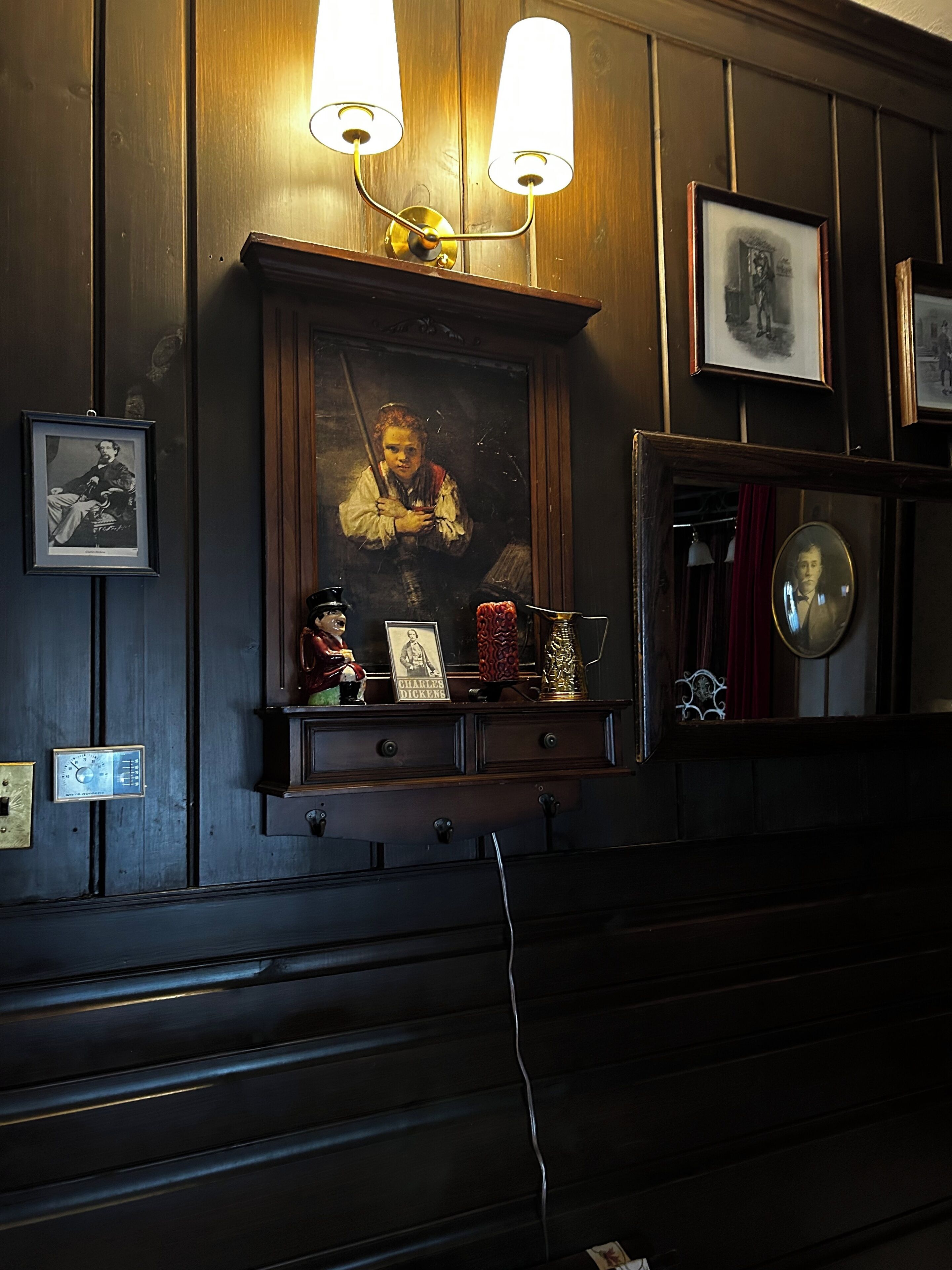 A room with a dark wooden wall, a framed portrait, a shelf with various items, and two wall-mounted lamps.