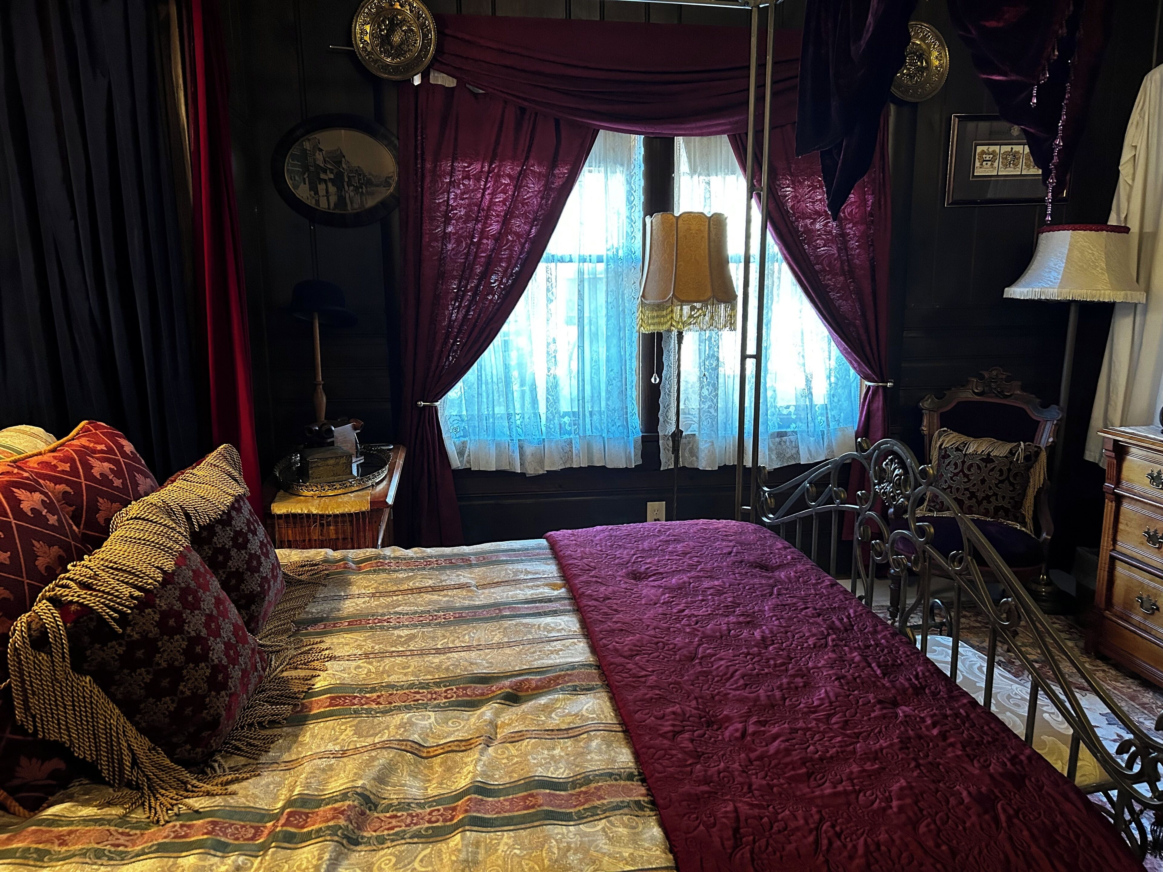 Ein Zimmer mit einem großen Bett, einem Schreibtisch und einem Stuhl. Ein Fenster mit Vorhängen und eine Lampe stehen auf dem Schreibtisch.