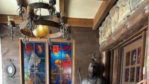 Un interior rústico con piso de madera, una mesa redonda con lámpara, una estatua y un ventanal de vidrio emplomado.