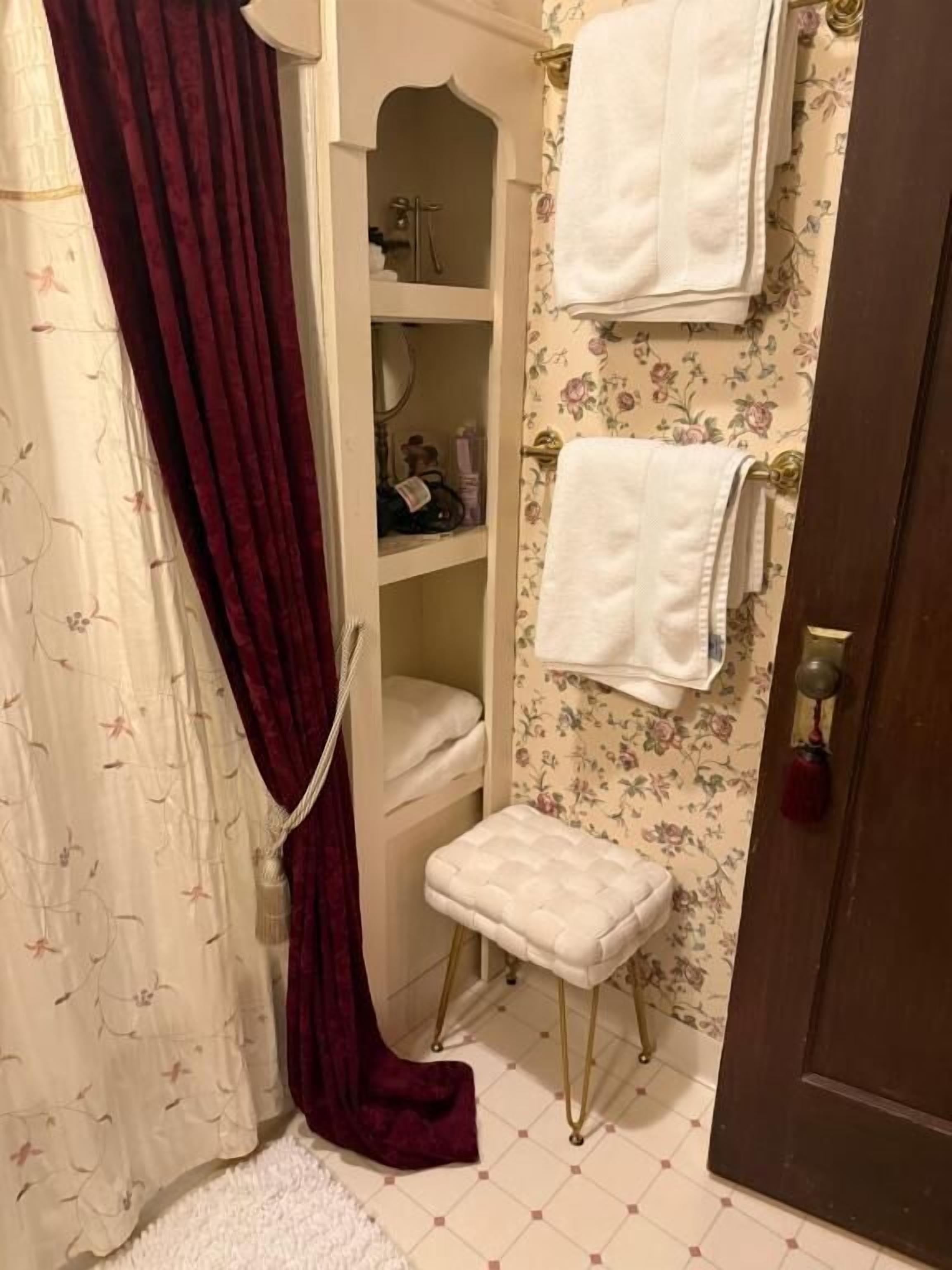 Un baño con tocador blanco, un taburete pequeño y un estante con artículos de aseo.