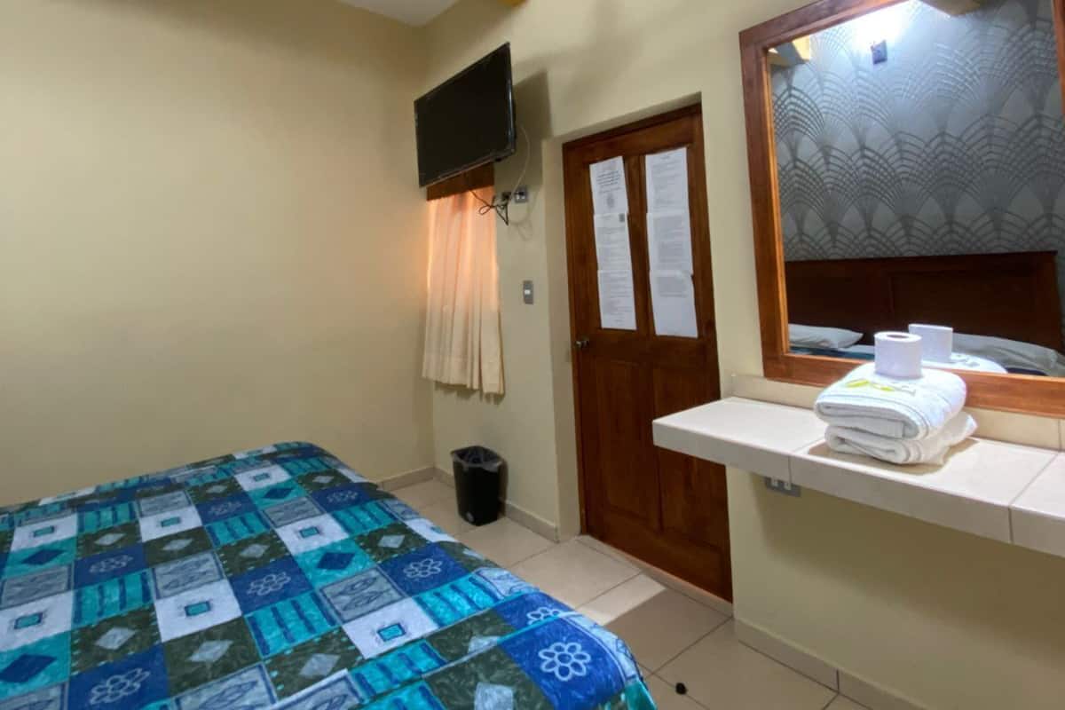A hotel room with a bed, a ceiling fan, a TV mounted on the wall, a bathroom mirror, and a towel rack.