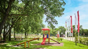 En lekplats med en gungbräda, en rutschkana och en klätterställning i en park med träd och flaggor.