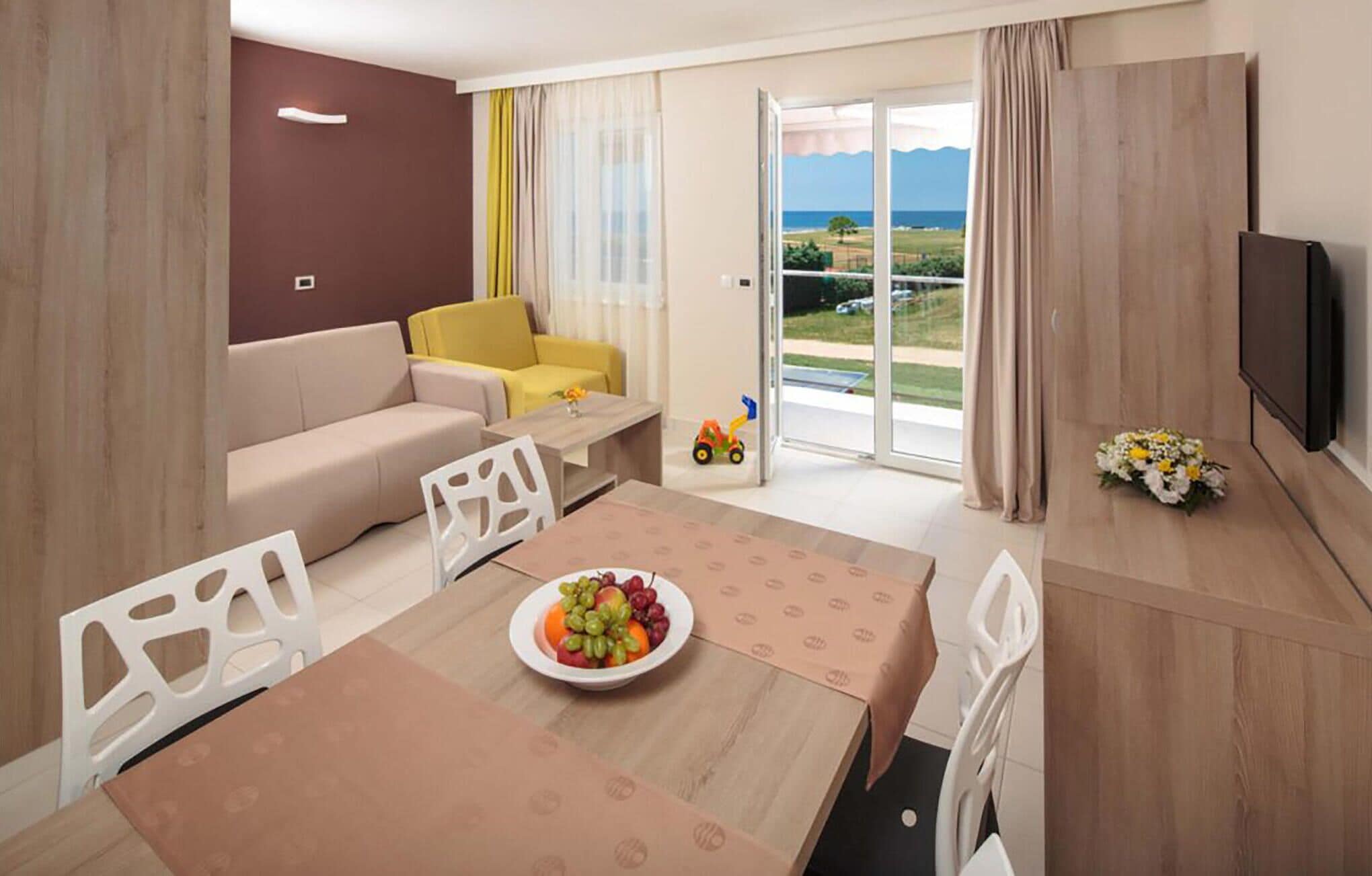 A modern living room with a sofa, a dining table with a fruit bowl, a TV, and a view of the outdoors.