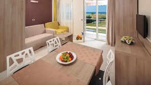 Ein modernes Wohnzimmer mit einer Couch, einem Esstisch mit Obstschale, einem Fernseher und Blick ins Freie.