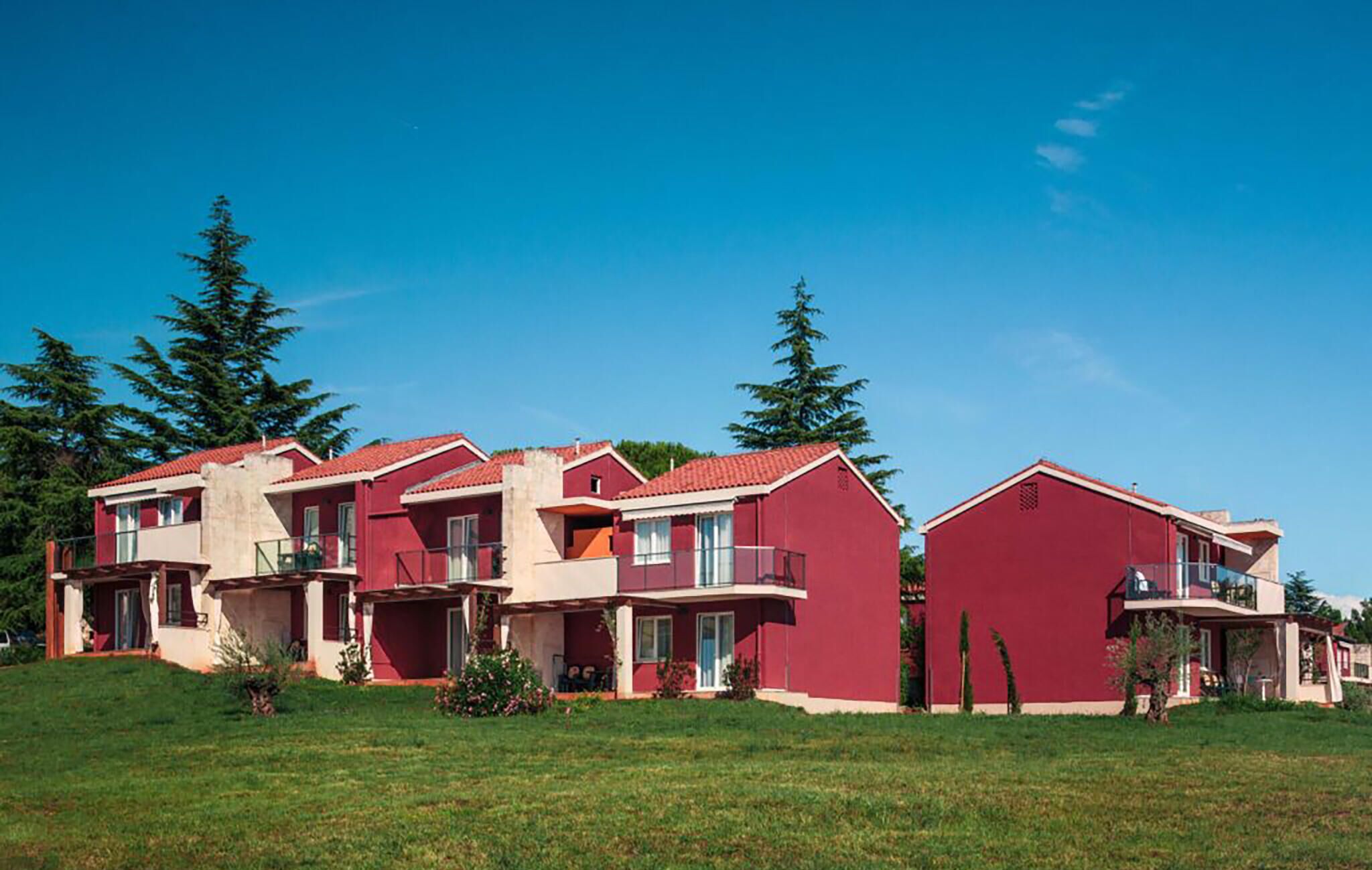 A row of red buildings with balconies, surrounded by green grass and trees.