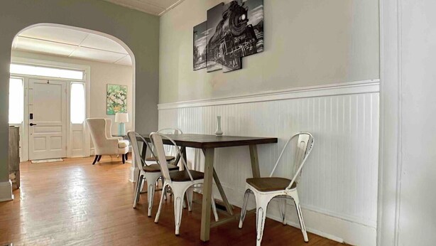A dining area with a wooden table and white chairs, a black and white painting on the wall, and a door leading to another room.