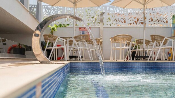 Ein Pool mit klarem Wasser, umgeben von weißen Stühlen und Sonnenschirmen, und ein weißer Zaun mit dekorativen Ausschnitten.