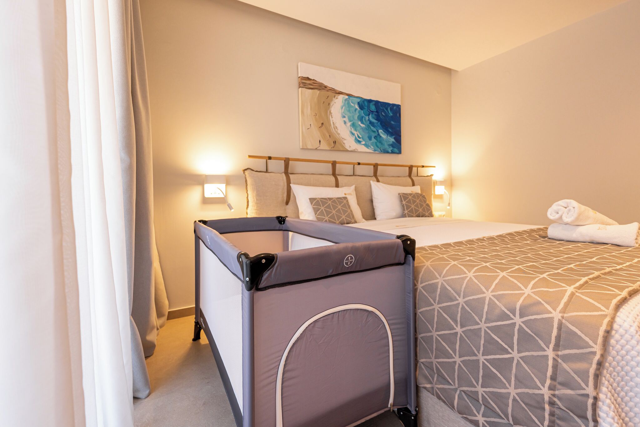 A hotel room with a bed, a headboard, a wall-mounted artwork, and a portable crib.