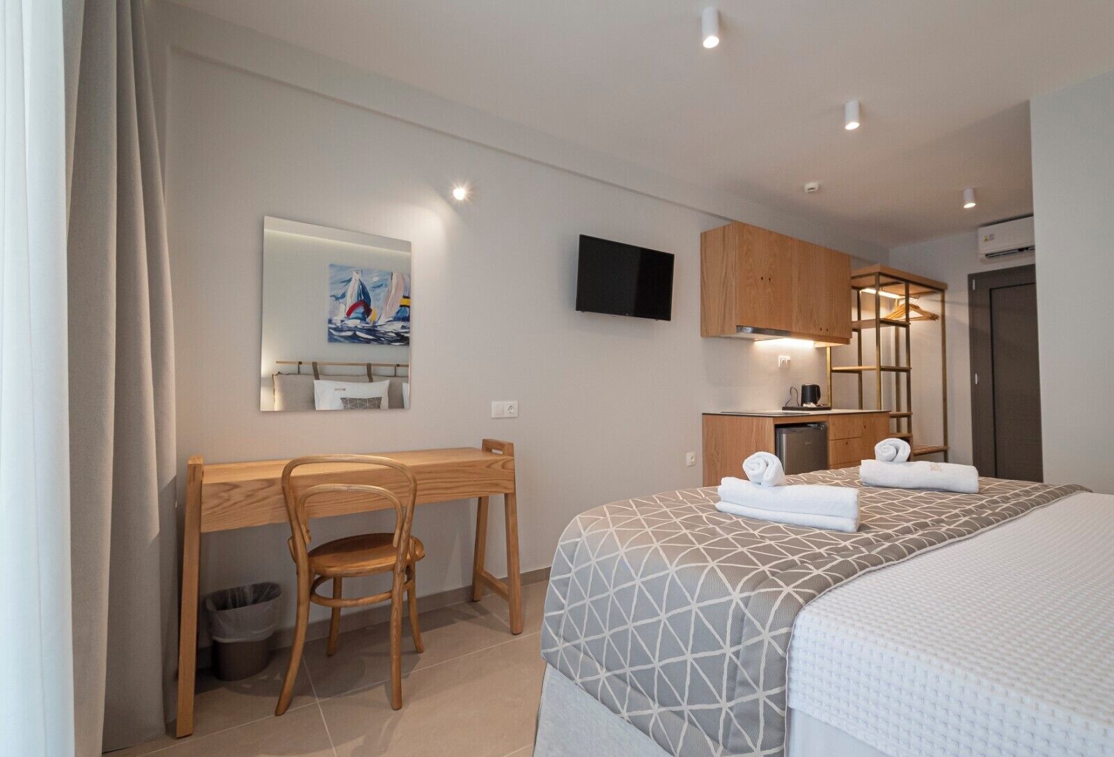 A hotel room with a bed, a desk, a chair, a TV, a mirror, a painting, and a kitchenette.