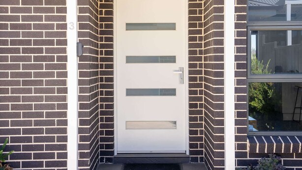 Una entrada de edificio moderna con una puerta blanca, paredes de ladrillo negro y una ventana a la derecha.