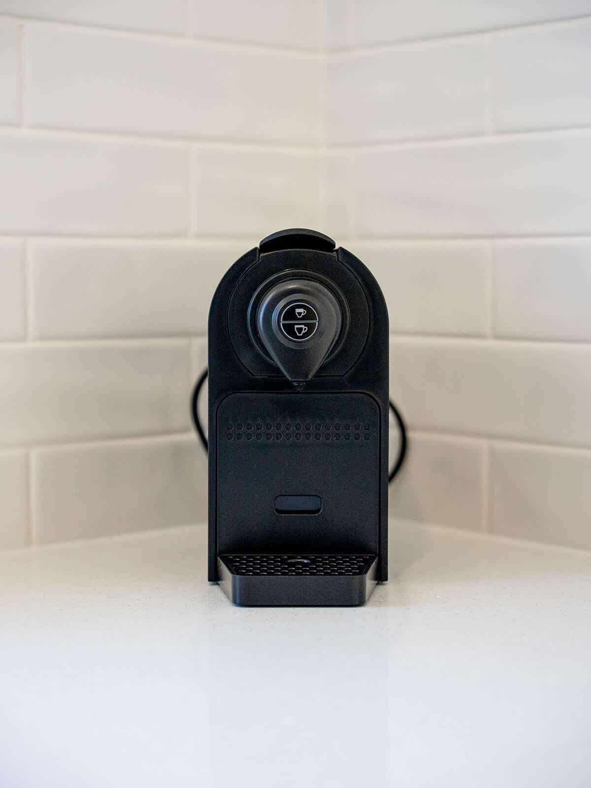 A black coffee machine with a drip tray, set against a white tiled wall.