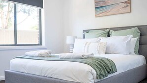 Una cama bien tendida con sábanas blancas y una manta verde, un cabecero tapizado en gris, y una pintura de una playa sobre ella.