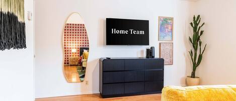 Un salon moderne avec un téléviseur à écran plat, un meuble de rangement noir, un canapé jaune, un grand miroir et une plante en pot.