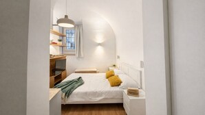 A compact bedroom with a bed, bedside tables, a hanging light fixture, a window with curtains, and a small shelf with items. - Studio Coppelle - Studio Apartment, Sleeps 2 (Rome)