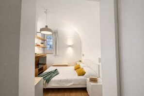 A compact bedroom with a bed, bedside tables, a hanging light fixture, a window with curtains, and a small shelf with items. - Studio Coppelle - Studio Apartment, Sleeps 2 (Rome)