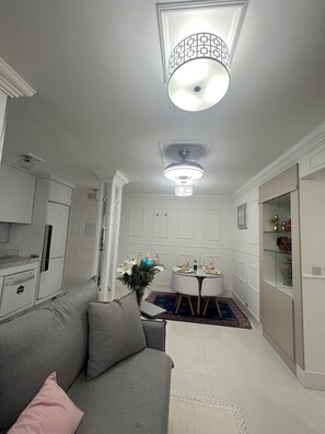 A modern living room with a grey sofa, a dining table with chairs, a ceiling fan, and a kitchen area with cabinets and a refrigerator. - Apto familiar fabuloso em condomínio/Interlagos, Zona Sul/IBC (Campo Grande)