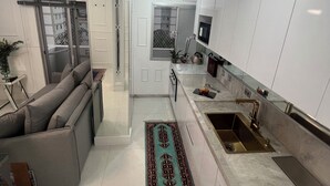 Eine moderne Küche mit weißen Schränken, einer Edelstahl-Spüle und einem Einbauofen. Im Wohnbereich befindet sich ein graues Sofa und ein gemustertes Teppich.