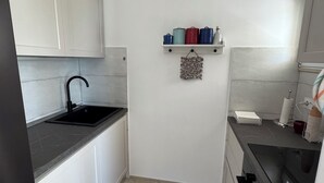 Et kompakt køkken med hvide skabe, en sort bordplade, en rustfri stålvask og en lille hylde med beholdere.