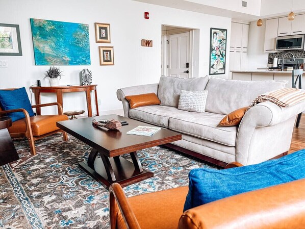 A modern living room with a sofa, coffee table, and chairs. There is a kitchen in the background.