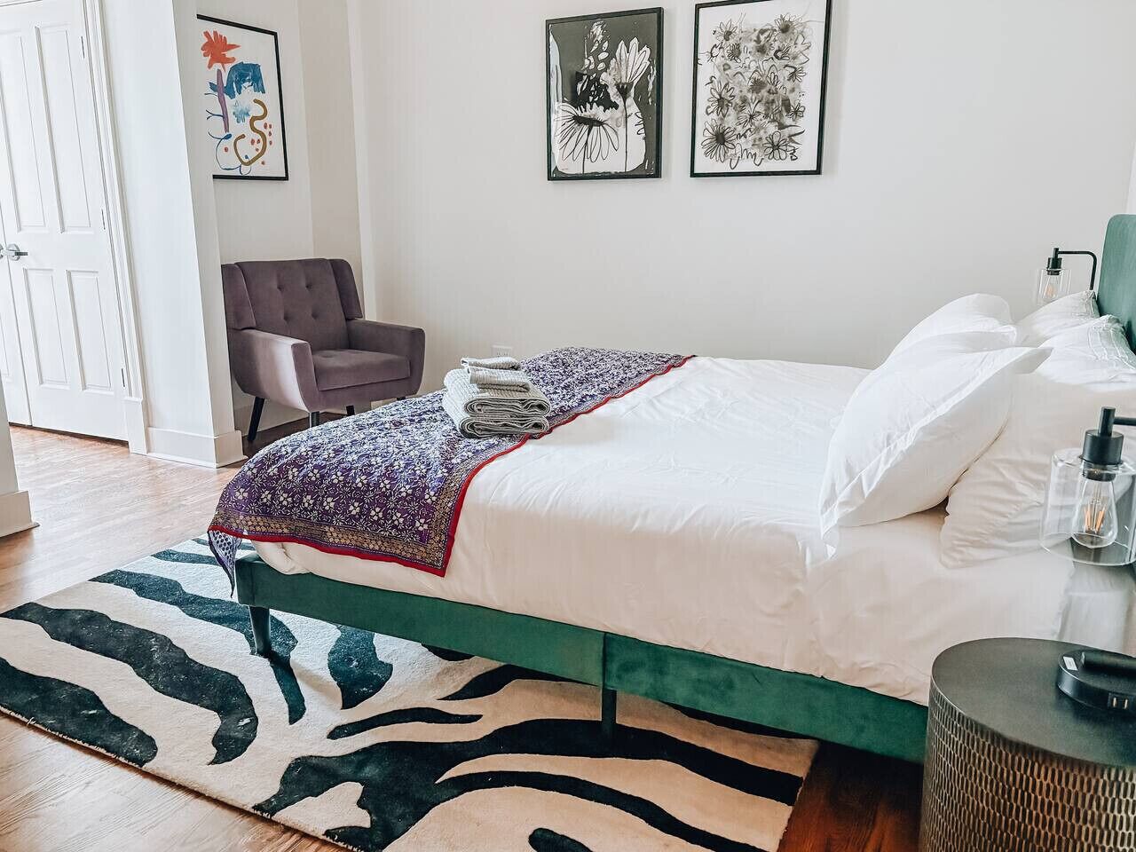 A bedroom with a green bed frame, white bedding, a zebra-patterned rug, and two framed floral artworks on the wall.