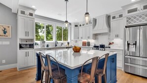Cozinha moderna com armários brancos, uma ilha grande com banquetas, eletrodomésticos de aço inoxidável e iluminação pendente.