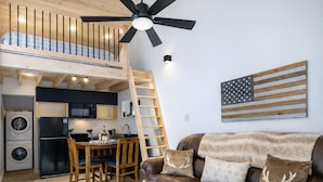 Eine moderne Loft-Wohnung mit Küche, Essbereich und Wohnzimmer. Sie verfügt über ein Schlafbett im oberen Bereich, einen Deckenventilator und eine große amerikanische Flagge als Dekoration.