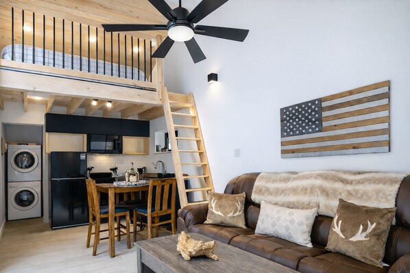 Eine moderne Loft-Wohnung mit Küche, Essbereich und Wohnzimmer. Sie verfügt über ein Schlafbett im oberen Bereich, einen Deckenventilator und eine große amerikanische Flagge als Dekoration.