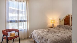 A bedroom with a bed, a chair, a nightstand with a lamp, and a window with sheer curtains.