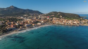 An aerial view of a coastal town with buildings along the shoreline, a mountain in the background, and clear blue waters.