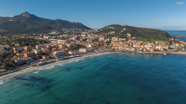Une vue aérienne d’une ville côtière, avec des bâtiments le long de la côte, une montagne en arrière-plan et des eaux d’un bleu limpide.
