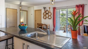 Une cuisine moderne avec un évier en acier inoxydable, un saladier bleu rempli de fruits, des chaises en bois et une vue sur la verdure à l’extérieur.