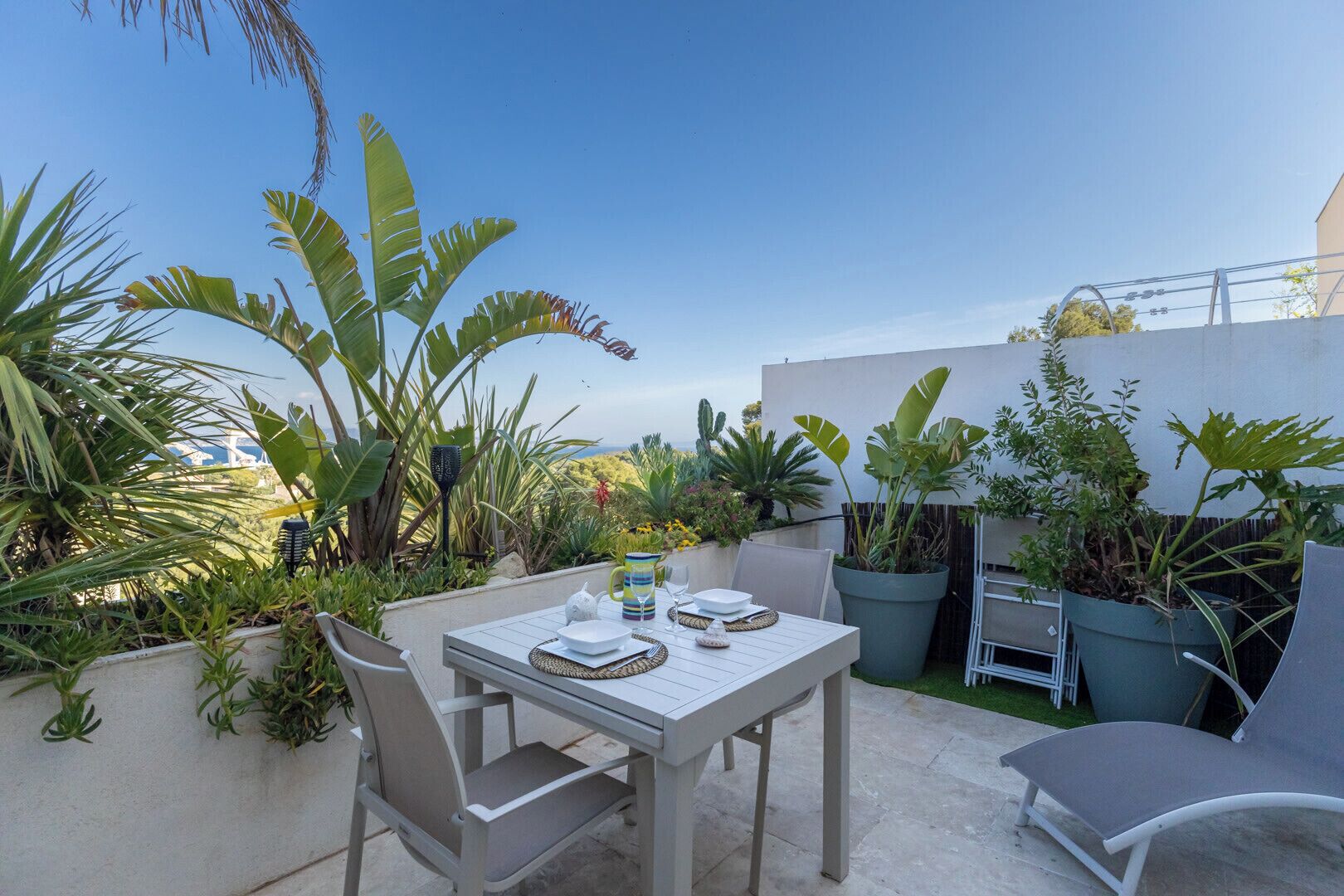 An outdoor patio with a table set for two, surrounded by potted plants and a clear sky.