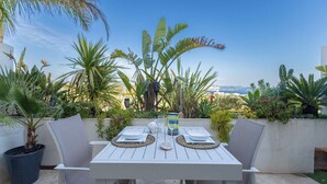 Eine Terrassengruppe im Freien mit einem weißen Tisch für zwei Personen, umgeben von verschiedenen tropischen Pflanzen und freiem Blick auf das Meer und entfernte Gebäude.