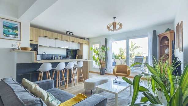 Ein modernes Wohnzimmer mit grauem Sofa, einem Couchtisch und einer Küchenzeile mit Holzschränken und einem Barhocker.